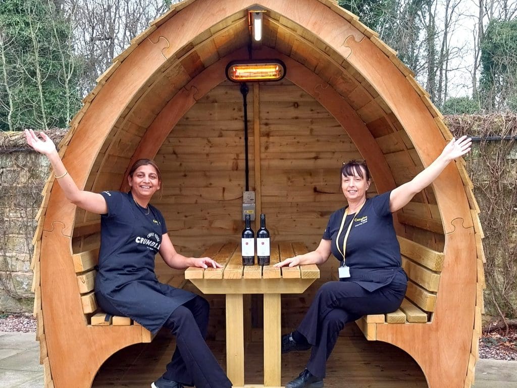 Two people sit in a heated outdoor pod with two bottles of wine, they smile at the camera