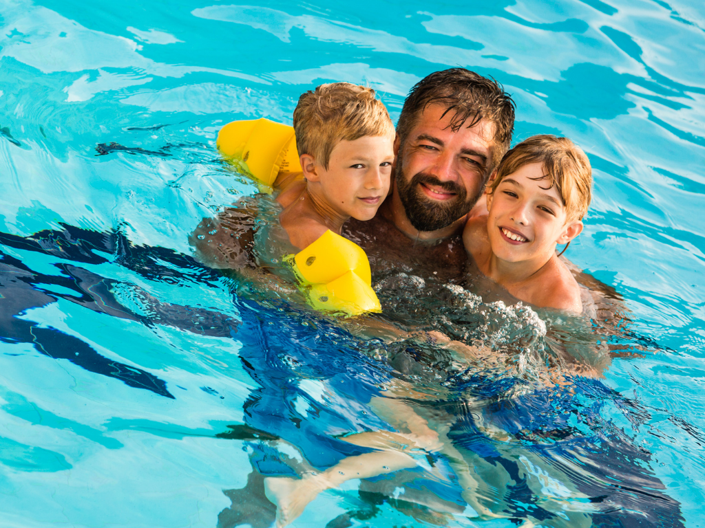 A parent and two children. one wearing armbands, are in a swimming pool smiling at the camera