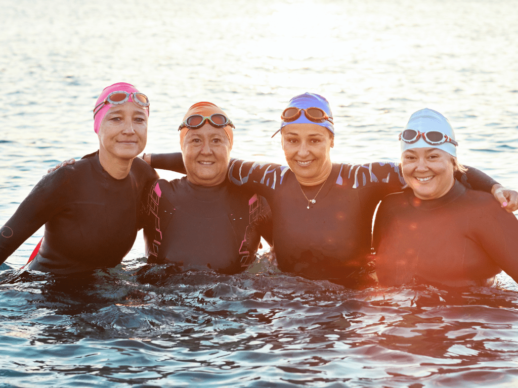 A group of women swimmers in the sea wearing wetsuits, swim caps and goggles. They have their arms around each other and are smiling at the camera